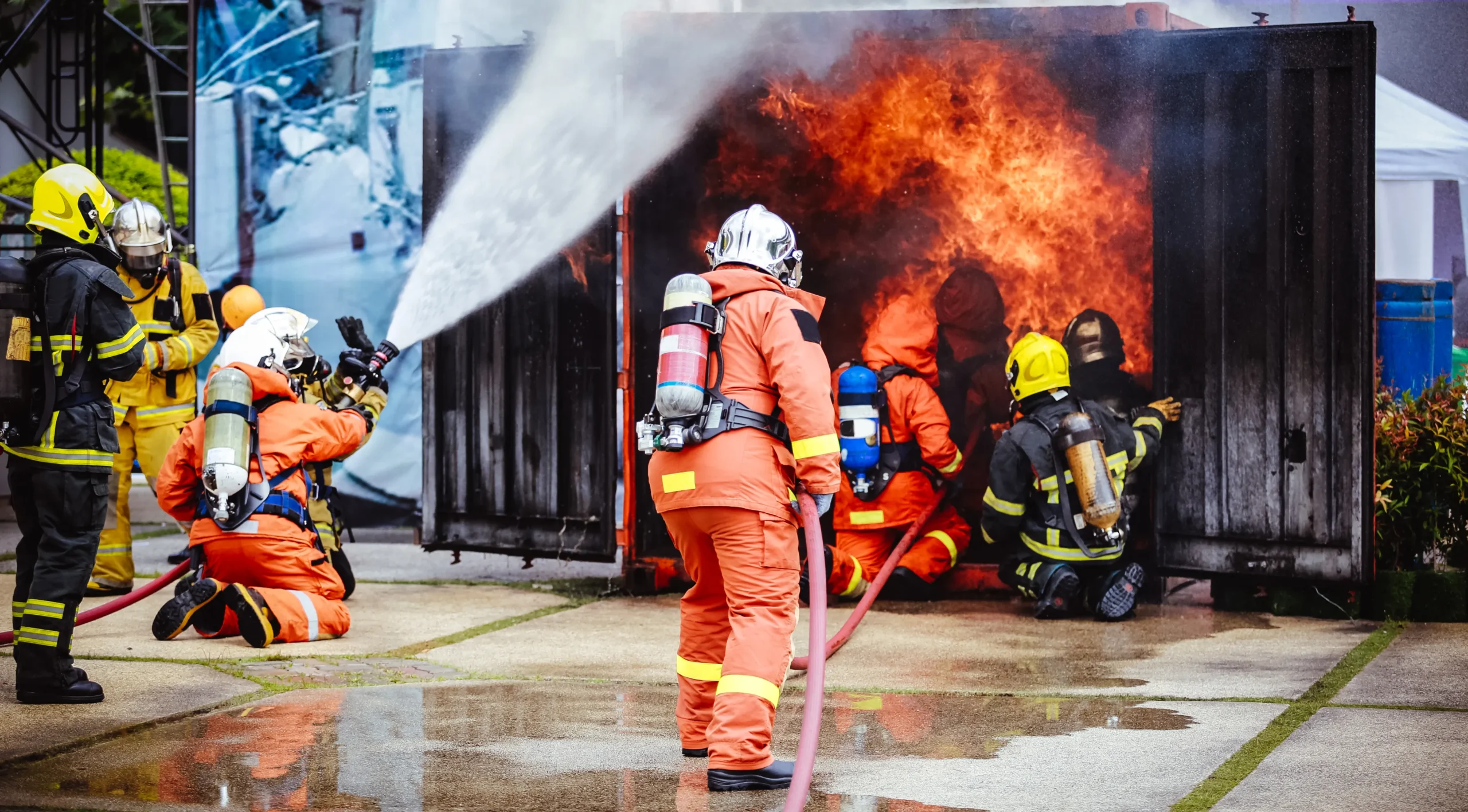 figters-against-fire-container_686e46df5b34b Firefighters in full gear actively extinguishing a blaze during a live fire training exercise outside a burning container.