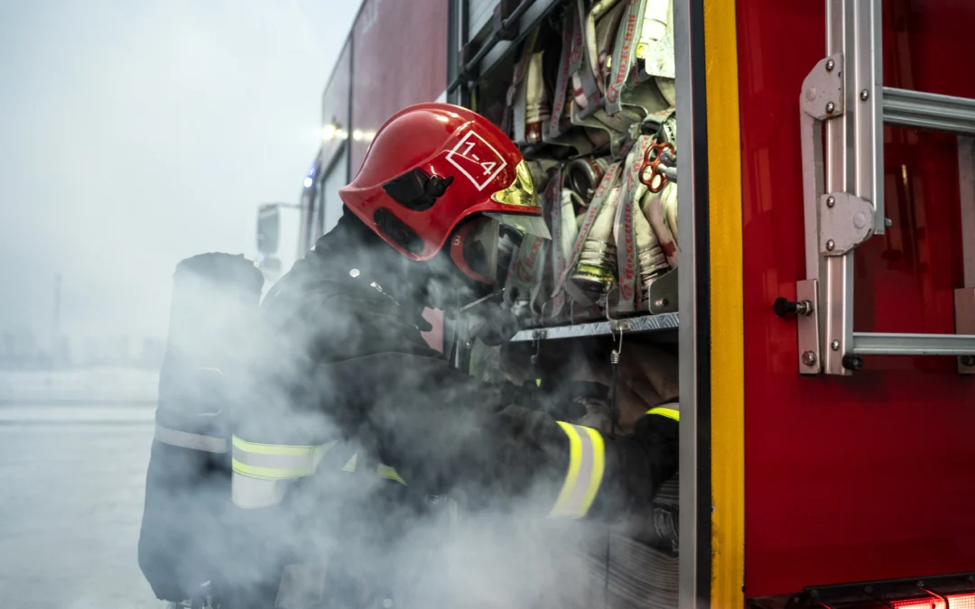 Firefighter in full gear and red helmet retrieving equipment from a fire truck amidst smoke.