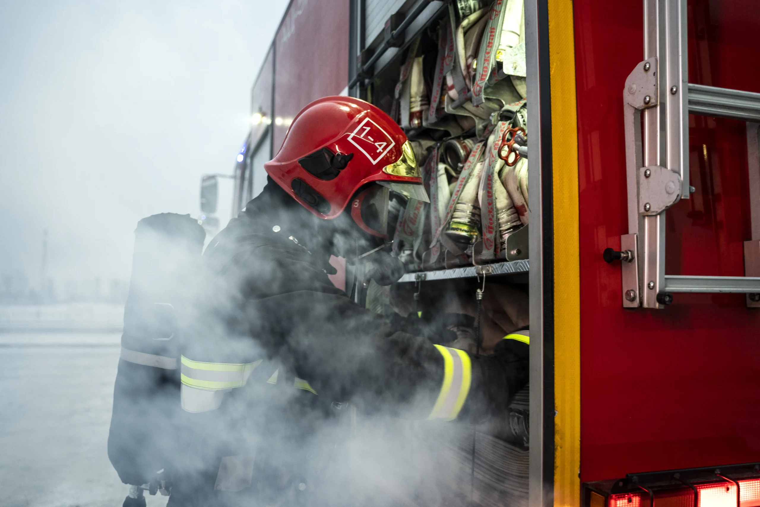 Firefighter in full gear and red helmet retrieving equipment from a fire truck amidst smoke.